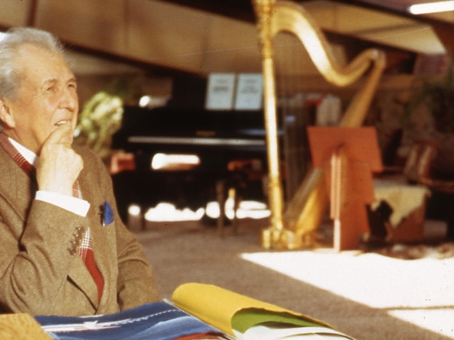 Frank Lloyd Wright working at his desk in the Garden Room at Taliesin West. Piano and harp in the background.