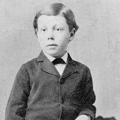 Black and white photograph of a young Frank Lloyd Wright, gazing directly at the camera. He is dressed in a dark Victorian-era suit with a buttoned-up jacket and a dark bow at his neck. His hair is neatly combed to the side.