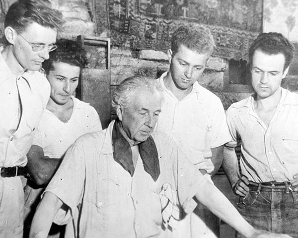 Black and white photo of Frank Lloyd Wright and a group of students gathered around a large table, examining architectural plans.