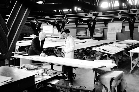 Black and white photo of two people working on a long drafting table in a spacious room with several other drafting tables and stools, exposed ceiling beams above, and papers and drafting tools scattered around the workspace.