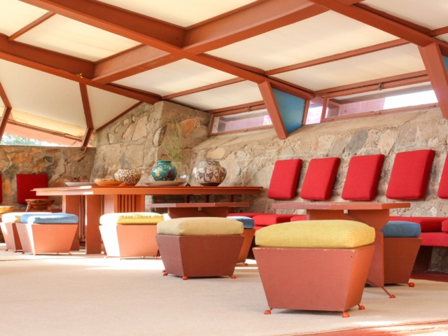 Interior view of Taliesin West showing a spacious room with stone walls and large windows under a vaulted ceiling. The room is furnished with tables and chairs in red and earth tones, and the seating includes cushioned stools and benches.