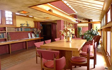 Interior of a Frank Lloyd Wright-designed residence, featuring horizontal lines, natural wood, and large tables with red chairs in an open-plan room illuminated by natural light.