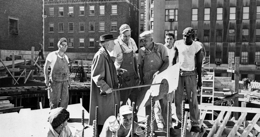 Black and white photograph of architect Frank Lloyd Wright wearing a fedora and overcoat, discussing plans with several construction workers on the site of the Guggenheim Museum.