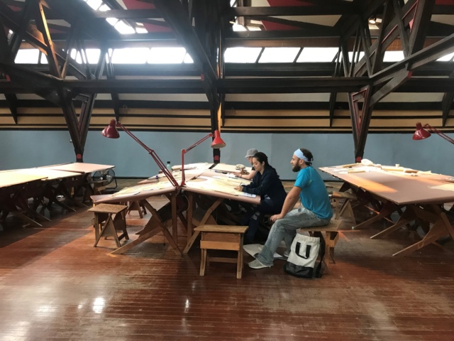 Interior of a spacious art studio with wooden floors and high ceilings featuring exposed beams. Natural light filters in through skylights. Three people are seated at long wooden tables with drawing boards, focused on their work.