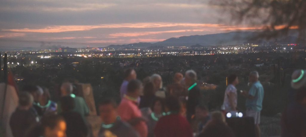 A group of people gathered at dusk, with glowing necklaces, engaging in conversation with a backdrop of a city's twinkling lights spread out beneath a twilight sky.