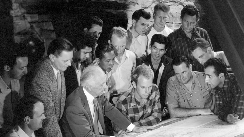 Black and white photo of Frank Lloyd Wright and a group of students gathered around a large table, examining architectural plans.