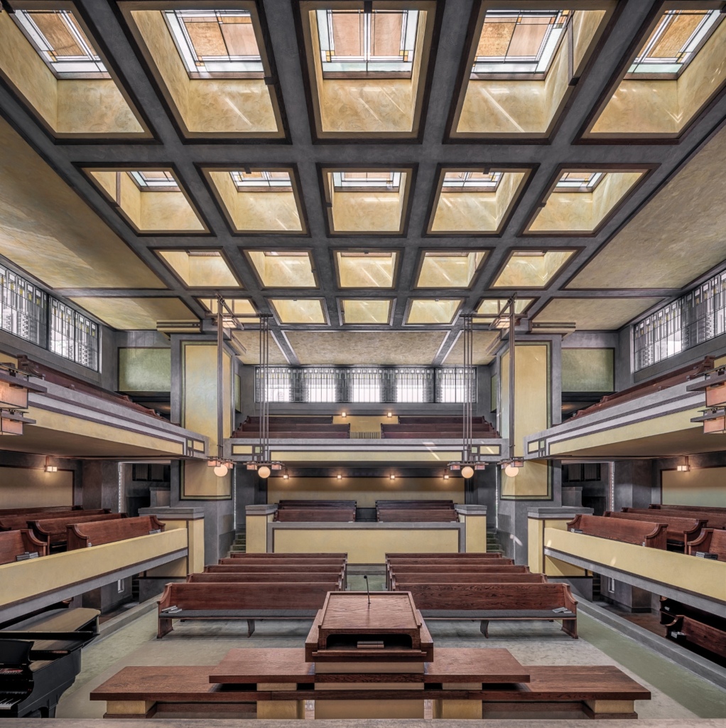 Interior view of Unity Temple showing the sanctuary with its distinctive architectural features. The room is characterized by strong horizontal lines, earthy tones, and geometric shapes.