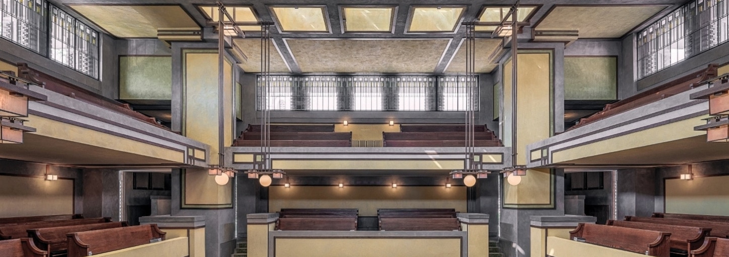 Interior view of Unity Temple showing the sanctuary with its distinctive architectural features. The room is characterized by strong horizontal lines, earthy tones, and geometric shapes.