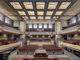 Interior view of Unity Temple showing the sanctuary with its distinctive architectural features. The room is characterized by strong horizontal lines, earthy tones, and geometric shapes.