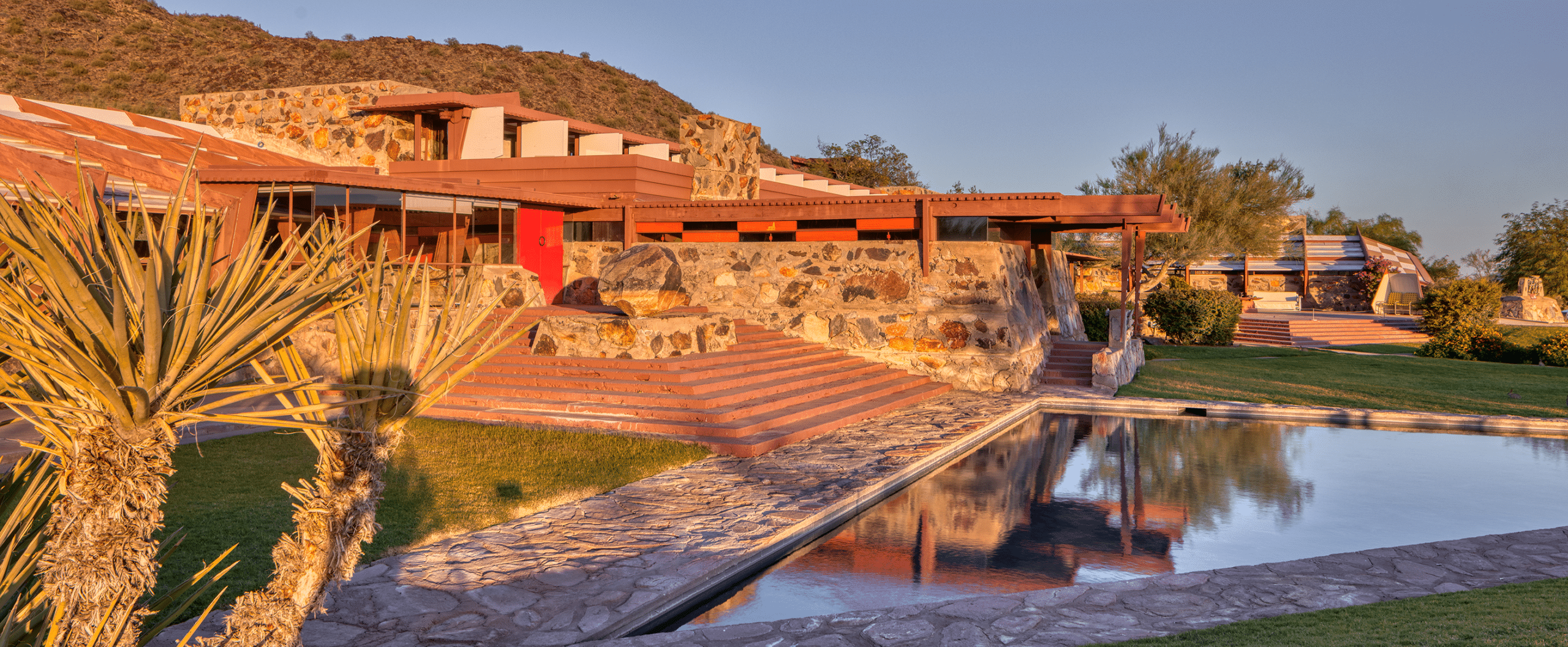 A view of a historic building on the property at Taliesin West made from desert rock material and concrete, with painted red wood doors and windows. There is a triangular pool in the foreground and a mountain with desert plant-life in the background. The image is framed by a bright blue sky.